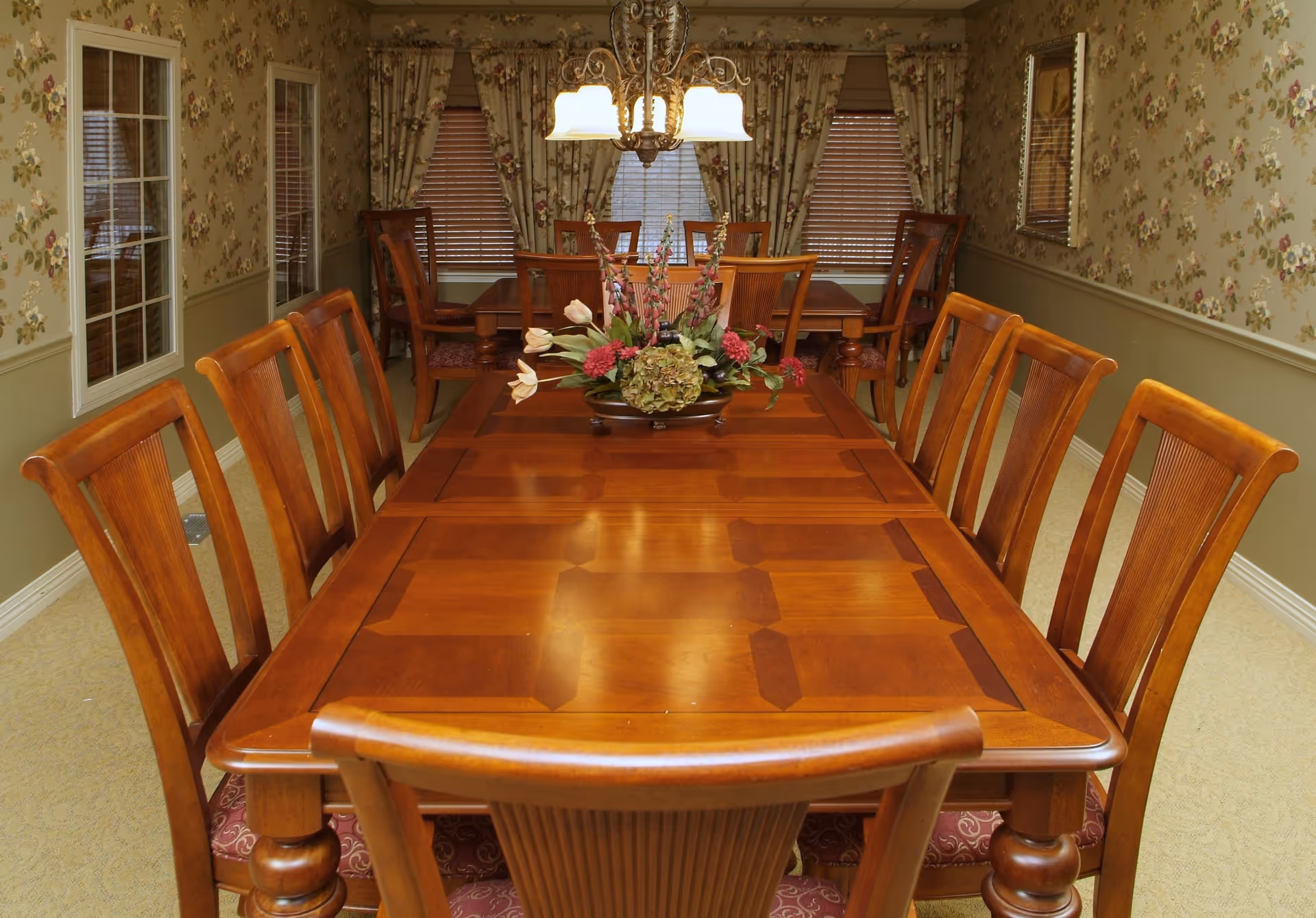 Long wooden dining table with chairs, a floral centerpiece, chandelier, and floral wallpaper in a dining room.