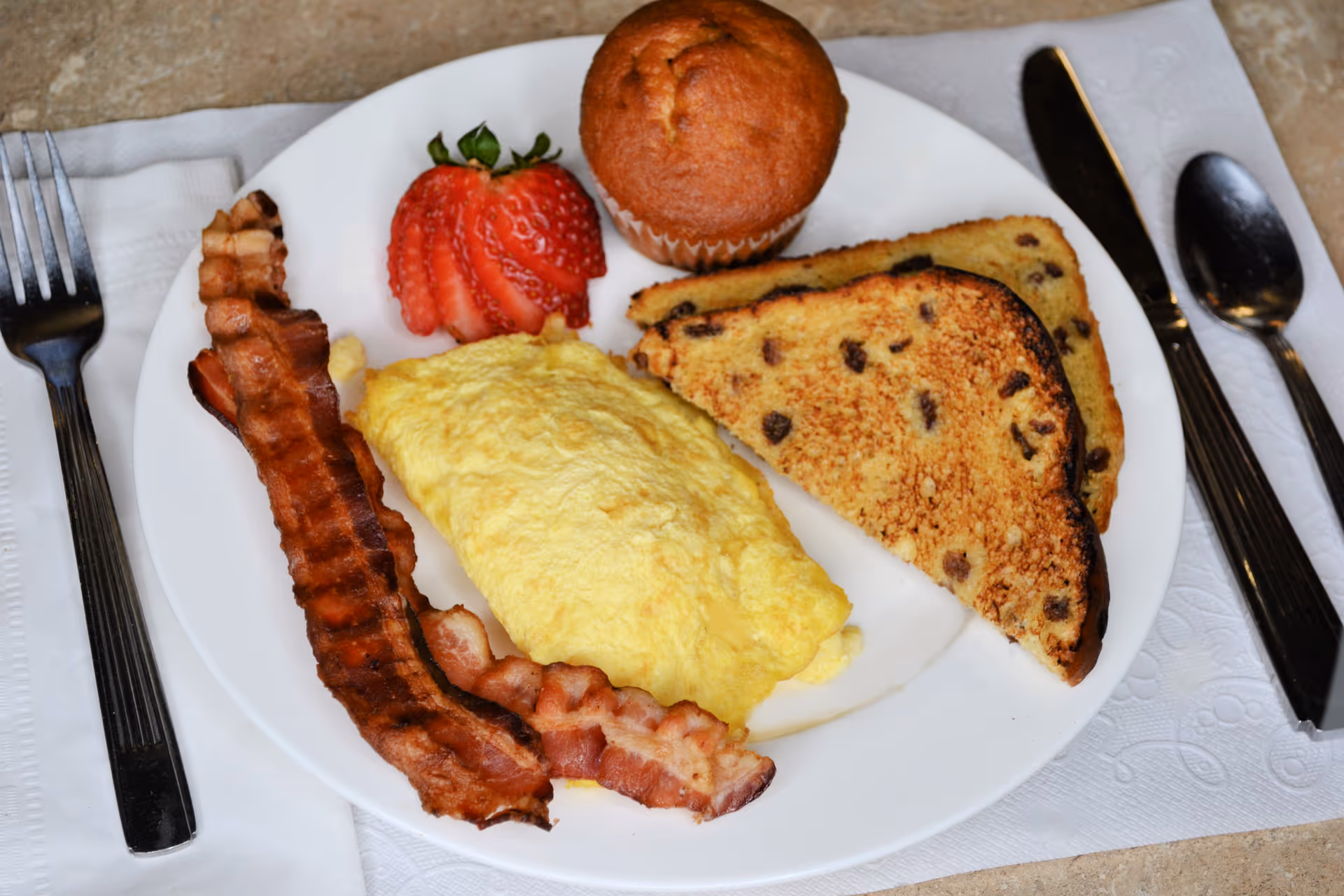 A breakfast plate with two strips of crispy bacon, a folded yellow omelette, two slices of toasted raisin bread, a sliced strawberry, and a muffin. The plate is set on a white napkin with a fork on the left and a knife and spoon on the right.