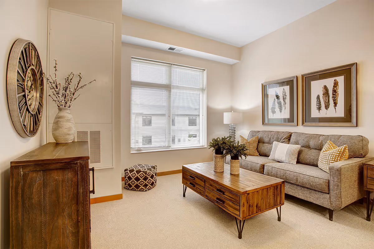 A cozy living room with a gray sofa adorned with patterned and striped pillows, a wooden coffee table with two decorative vases containing plants, a large window with blinds, a wooden sideboard with a large round wall clock and a vase with branches, and two framed botanical prints on the wall.