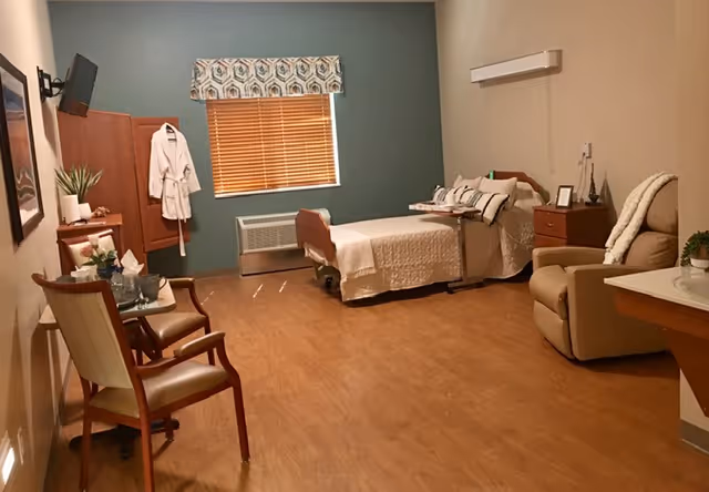 A spacious senior living bedroom with a single bed covered in a beige quilt, a reclining chair with a blanket draped over it, a wooden nightstand with decorative items, a small table with two chairs, a wall-mounted TV, a window with wooden blinds and a patterned valance, and a wooden floor.