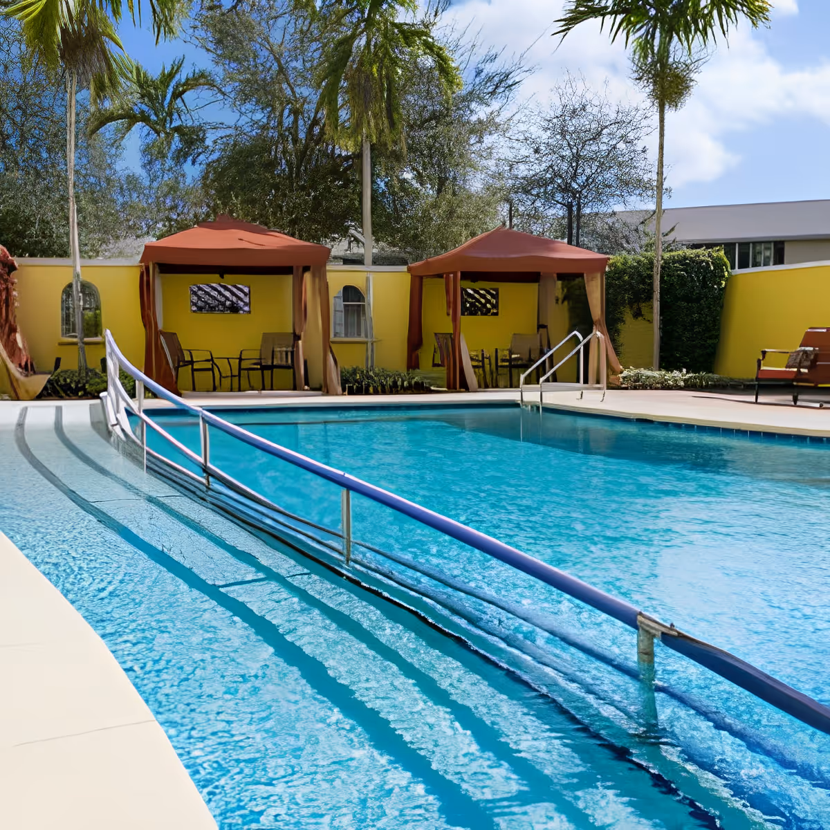 Outdoor swimming pool with a handrail ramp leading into the water, surrounded by a yellow wall. Two red canopy tents with chairs underneath are visible in the background, along with palm trees and other greenery under a partly cloudy sky.