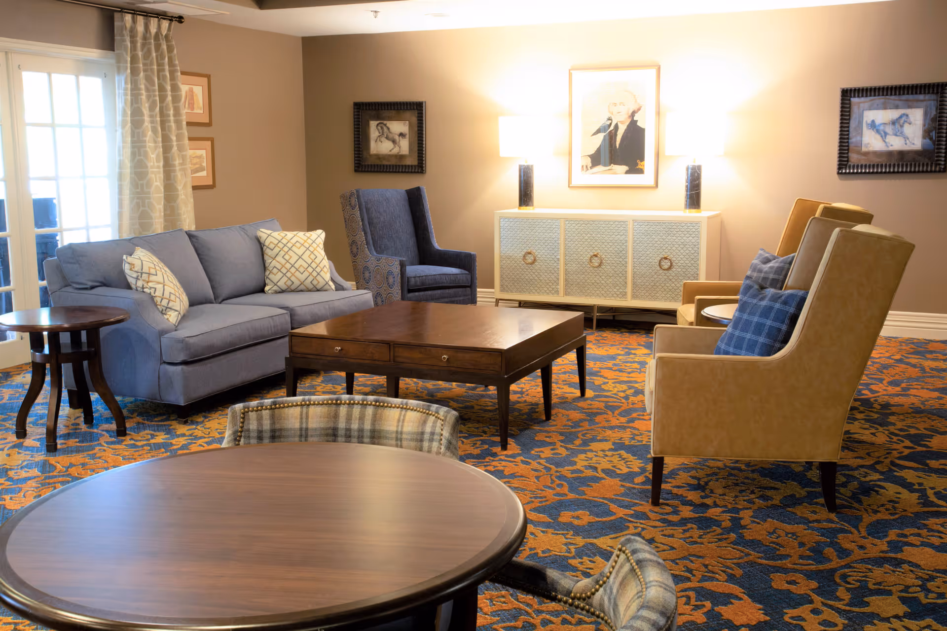 A cozy living room area with a blue sofa, two beige armchairs with blue plaid pillows, a blue patterned armchair, and a wooden coffee table in the center. There is a round wooden table with plaid chairs in the foreground. The room has a patterned carpet in blue and orange tones, beige walls with framed artwork, and a sideboard with two lamps and a portrait above it.