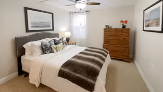 A neatly made bed with white linens, multiple decorative pillows, and a brown throw blanket in a bedroom. The room features a wooden dresser with a vase of red flowers and framed photos on top, a ceiling fan with light, two framed pictures on the walls, and a window with a sheer curtain allowing natural light.