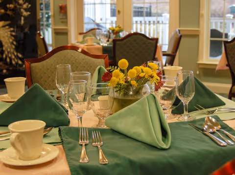 A formal dining table set with green napkins, glassware, silverware, and a small yellow floral centerpiece in a dining room.