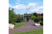 A paved pathway in a garden area leading to a wooden gazebo surrounded by green bushes and trees under a partly cloudy sky.