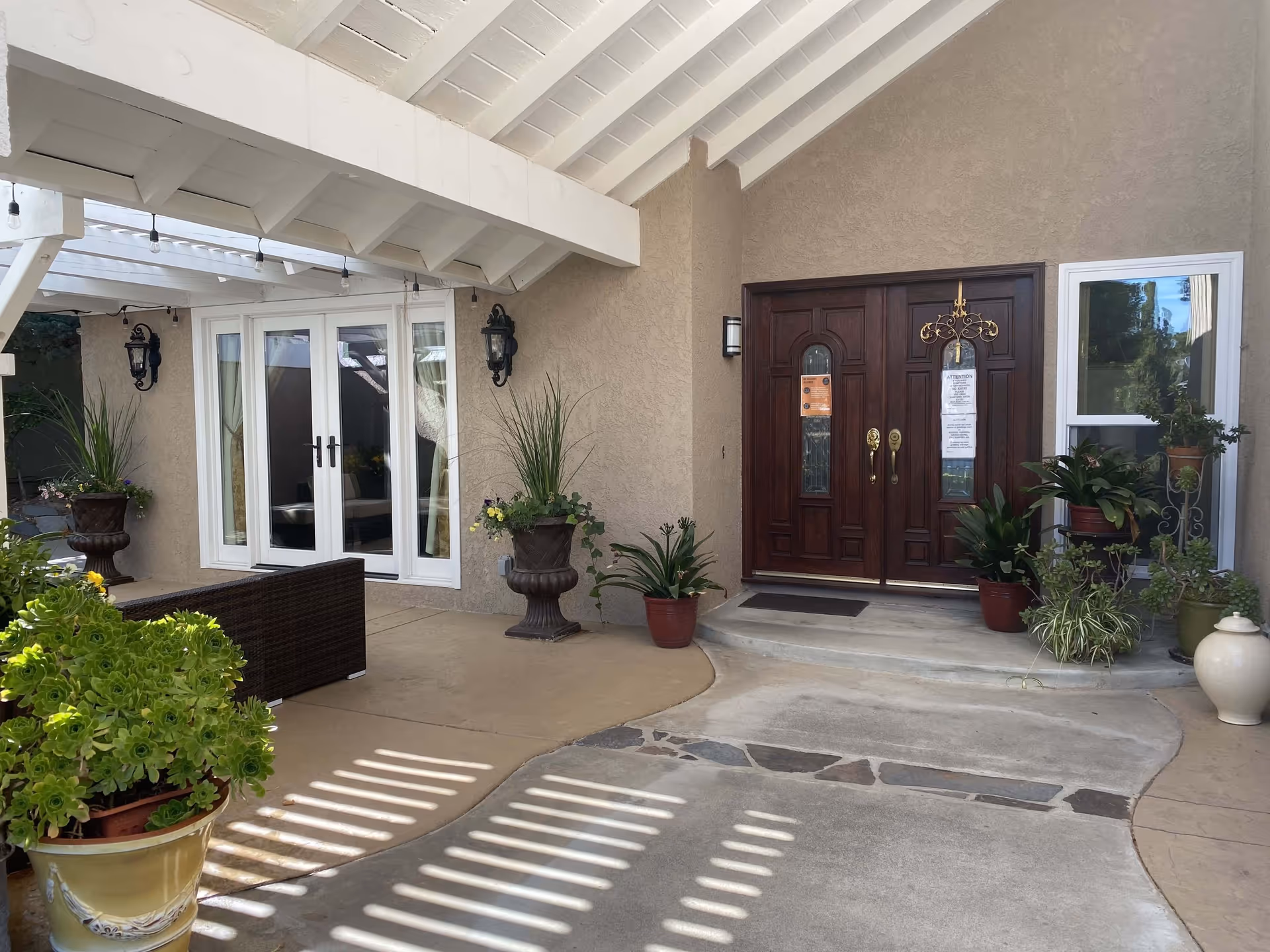 Covered outdoor entrance area of a building with a wooden double door, potted plants, and a seating area with a wicker bench. The ceiling is white with exposed beams and hanging string lights. There are two wall-mounted lantern-style lights on either side of the doors.