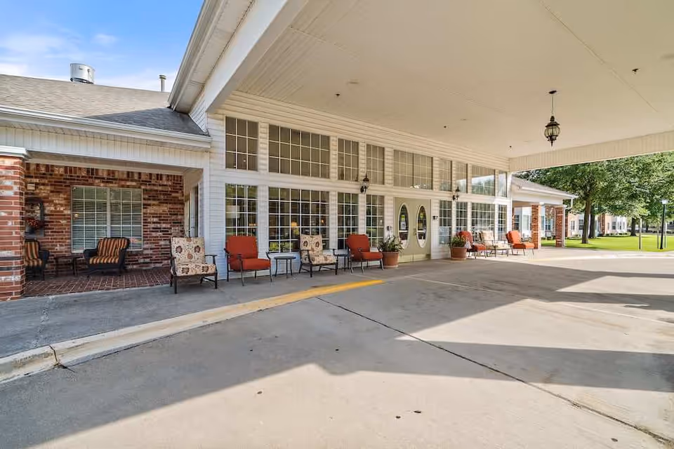 Covered entrance area of Morada Grand Prairie facility with several cushioned chairs and small tables arranged along the wall with large windows and double doors. The exterior features brick and white siding with hanging lantern-style lights and a concrete driveway.