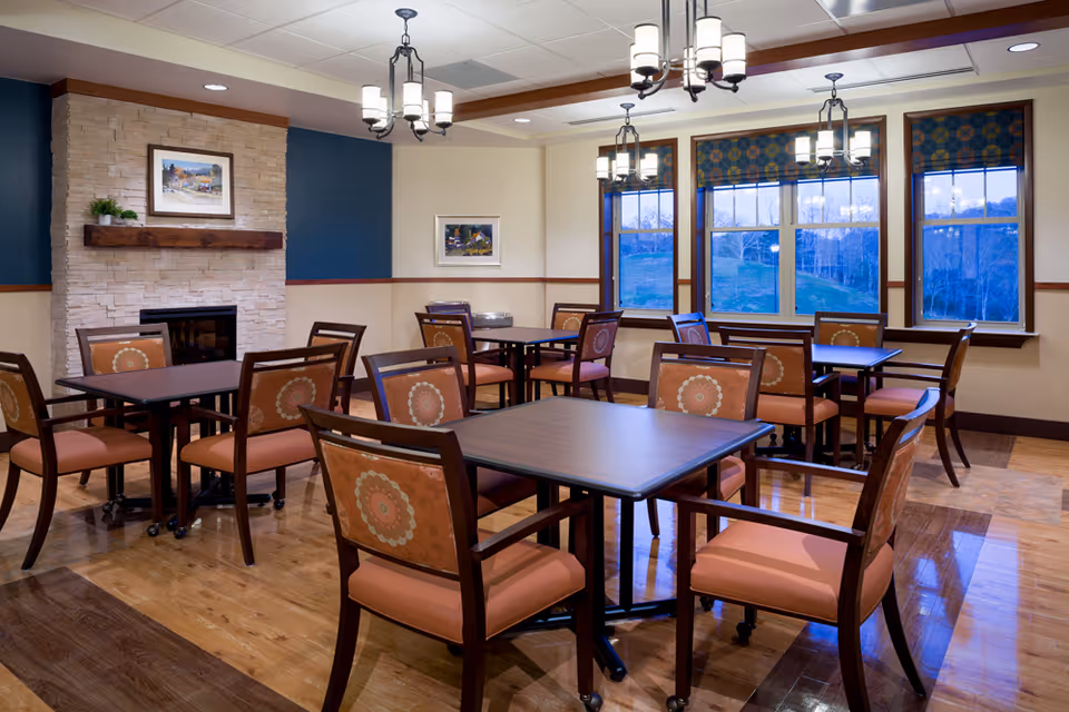 A dining room with several square tables and cushioned chairs featuring a floral pattern on the backrests. The room has wooden flooring, a stone fireplace with a wooden mantle, framed artwork on the walls, and large windows with patterned valances showing an outdoor view. Ceiling lights hang above the tables providing warm lighting.