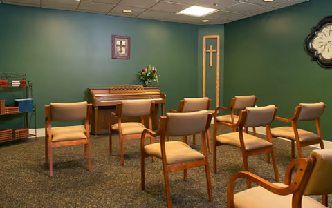 A small room with green walls featuring a piano against the back wall, a framed cross picture above it, and a wooden cross decoration on the right wall. Several wooden chairs with beige cushions are arranged facing the piano. There is a clock on the right wall and a small shelving unit on the left side of the room.