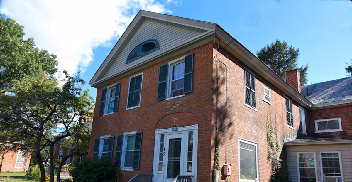 Two-story red brick house with green shutters, a central white entrance, and trees in the yard under a blue sky.