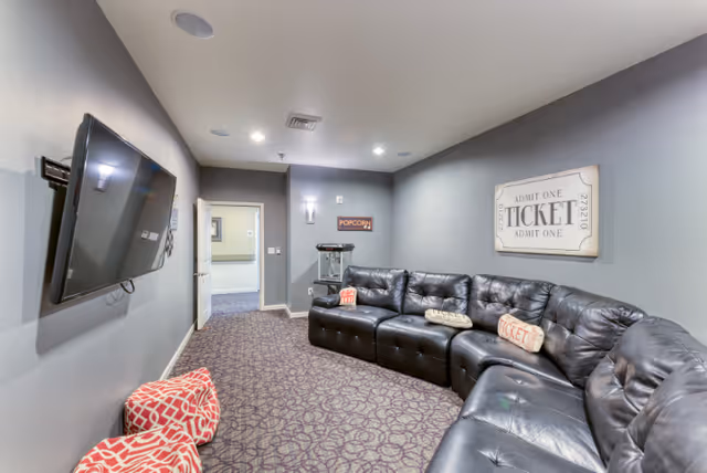 A cozy entertainment room with a large black leather sectional sofa adorned with decorative pillows, a wall-mounted flat-screen TV, a popcorn machine in the corner, and a sign on the wall that reads 'ADMIT ONE TICKET'. The room has gray walls and patterned carpet flooring.