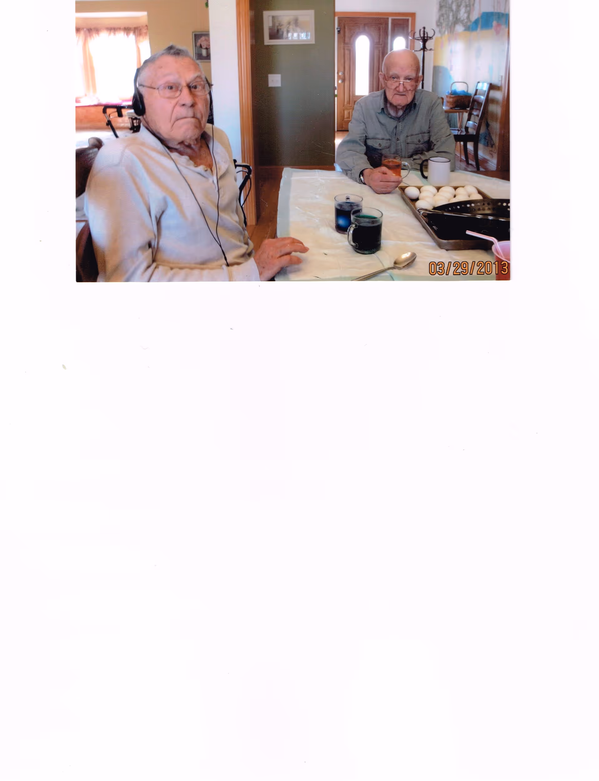 Two elderly men sit at a dining table with mugs and a tray of eggs in a home interior.