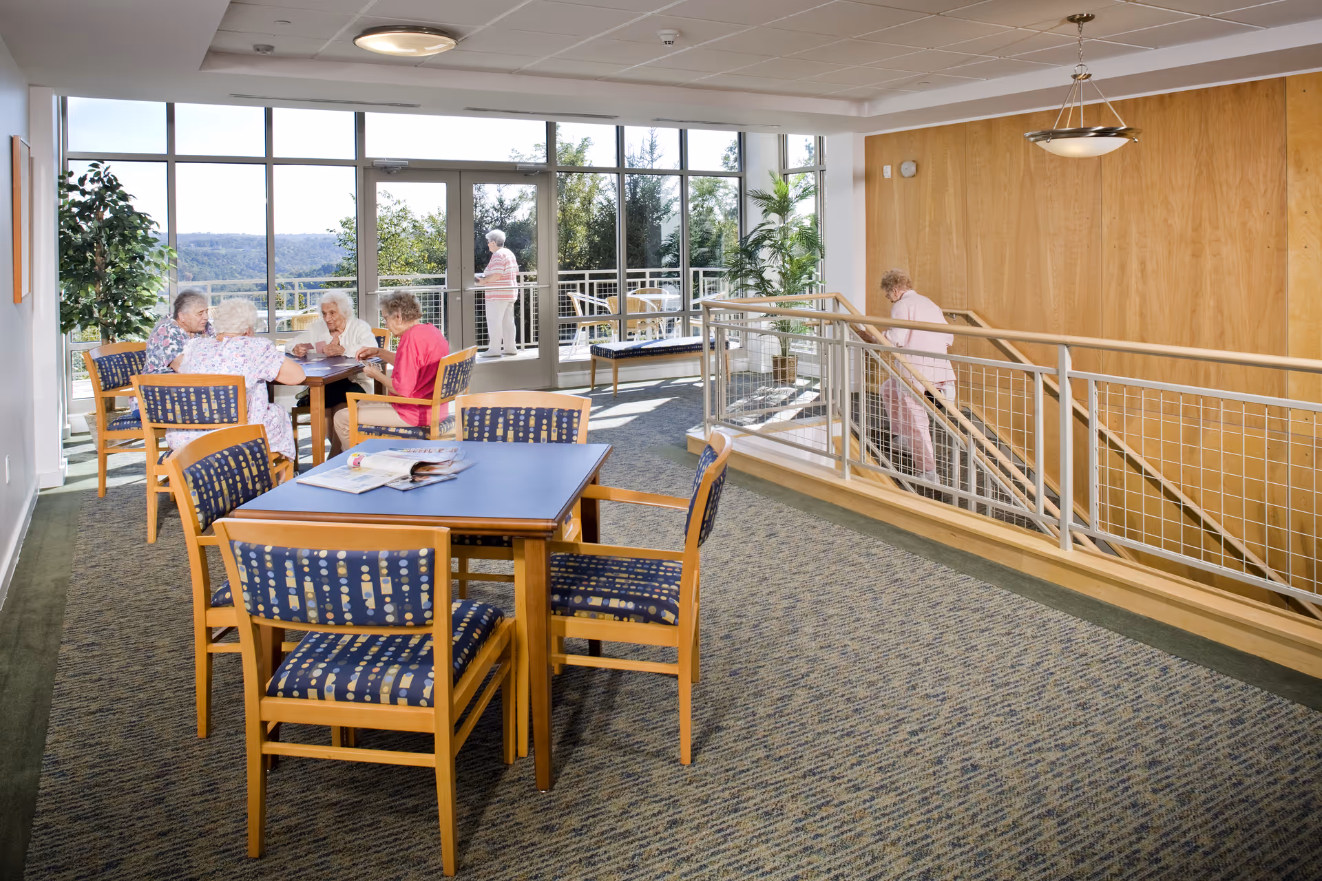 A bright common area in a retirement residence with large windows showing an outdoor view. Several elderly women are seated around a table playing a game, while one woman stands outside on a balcony and another is walking up a staircase. The room has carpeted floors, wooden paneling on one wall, and wooden tables and chairs with patterned cushions.
