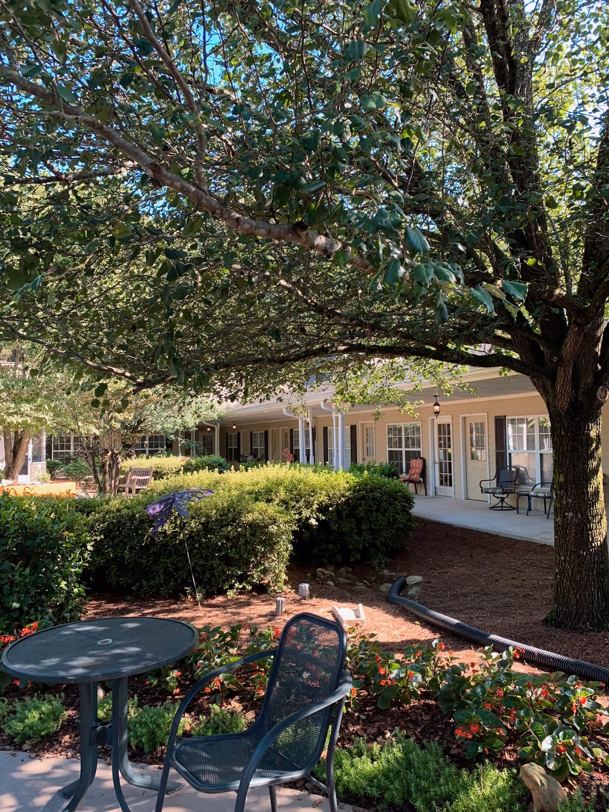 Outdoor garden area at Sunabella of Towne Lake with a round metal table and chair in the foreground, surrounded by green bushes, flowers, and a large tree providing shade. In the background, there is a covered porch with chairs and windows of the facility building visible.