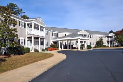 Exterior view of a two-story senior living facility building with gray siding and white trim. The building features balconies, a covered entrance, and a paved driveway with a sidewalk and landscaped greenery including trees and bushes under a clear blue sky.