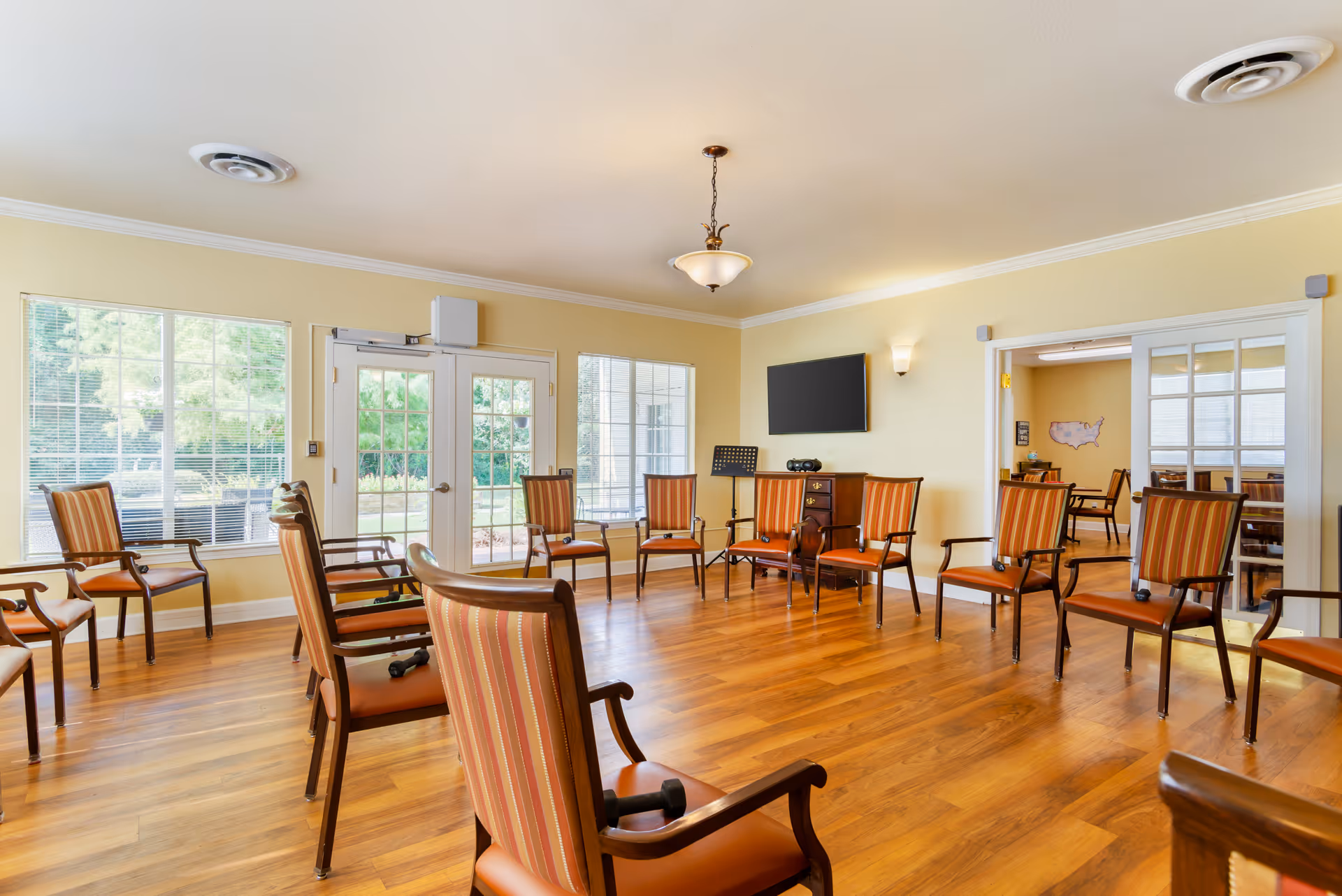 A bright room with wooden flooring and beige walls, arranged with multiple wooden chairs with orange striped cushions placed in a circle. There are windows and glass doors letting in natural light, a wall-mounted TV, a small cabinet, and a music stand in the room. Another room with similar chairs is visible through glass double doors.