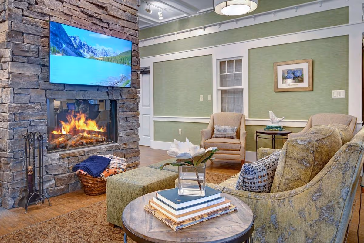 Cozy living room area with a stone fireplace that has a fire burning inside. Above the fireplace is a large flat-screen TV displaying a scenic mountain and lake view. The room features green walls with white trim, a comfortable armchair, a sofa with patterned pillows, a round wooden coffee table with a vase holding a white flower, and a basket with folded blankets on the floor.