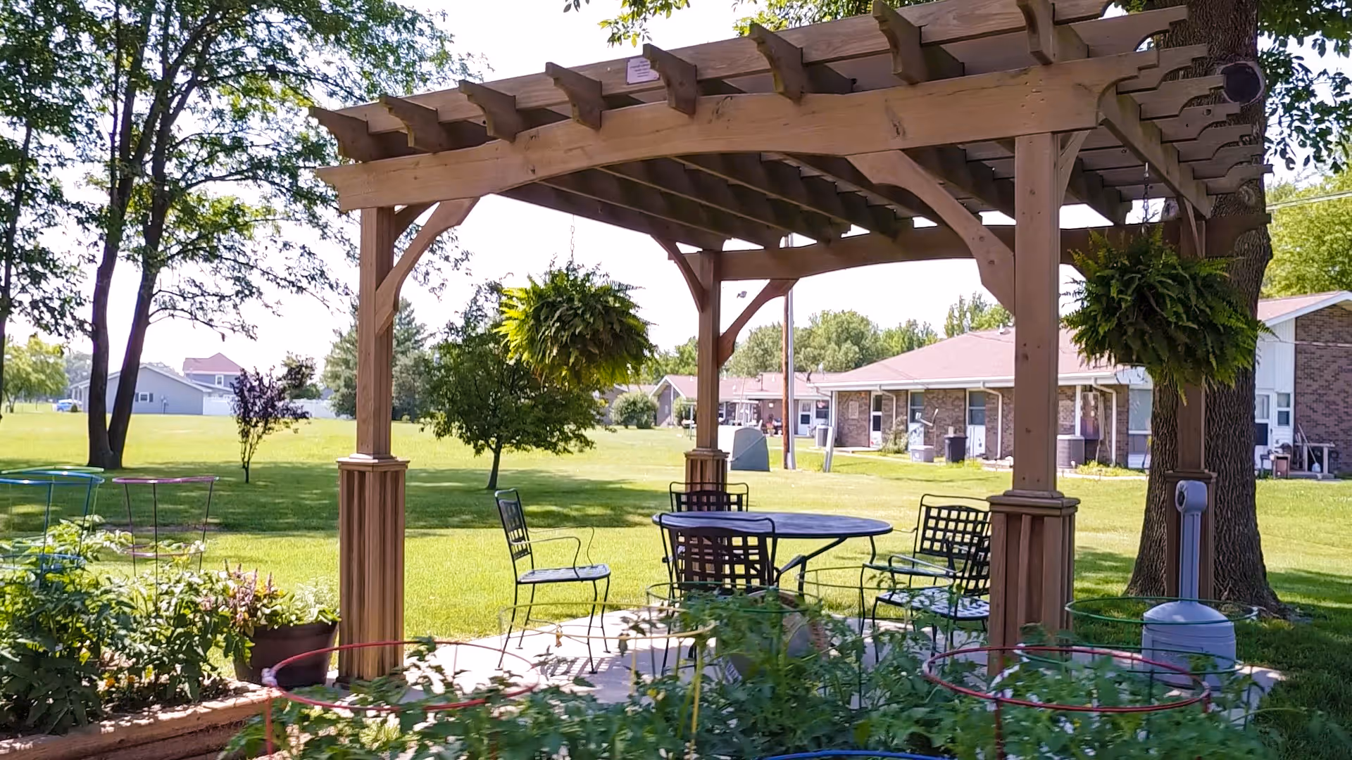 Outdoor seating area under a wooden pergola with hanging plants, metal chairs, and a round table on a concrete patio surrounded by green grass and trees, with single-story brick buildings in the background.