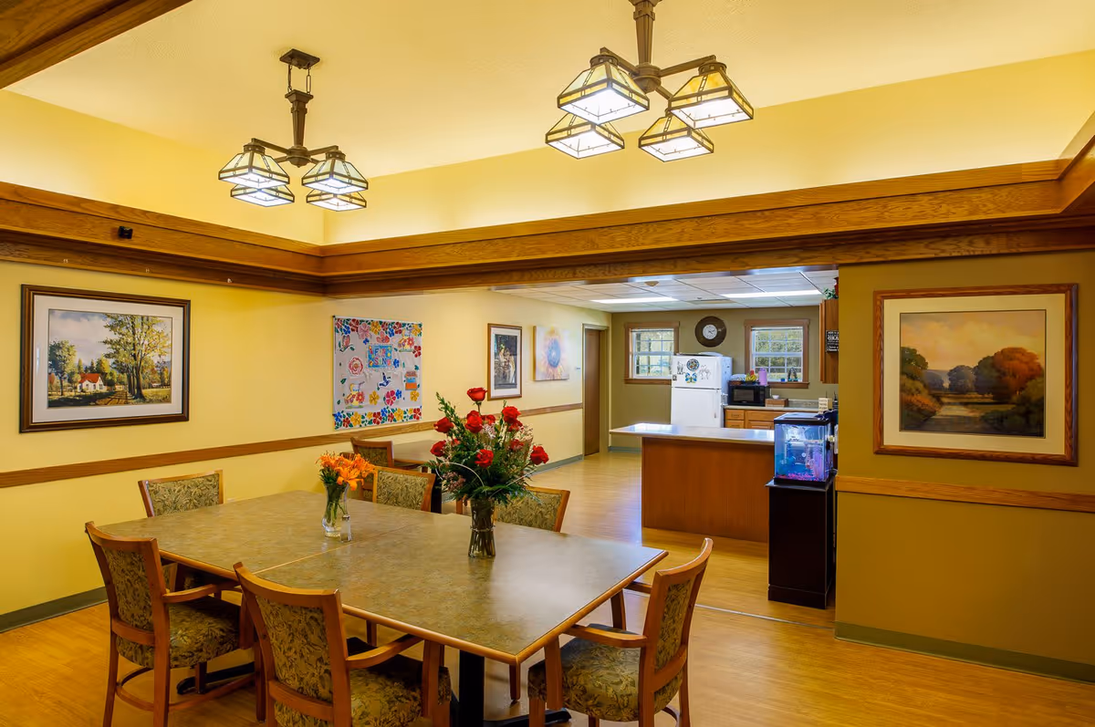 Well-lit dining room with a table and chairs, floral centerpieces, framed artwork, and a kitchenette visible in the background.