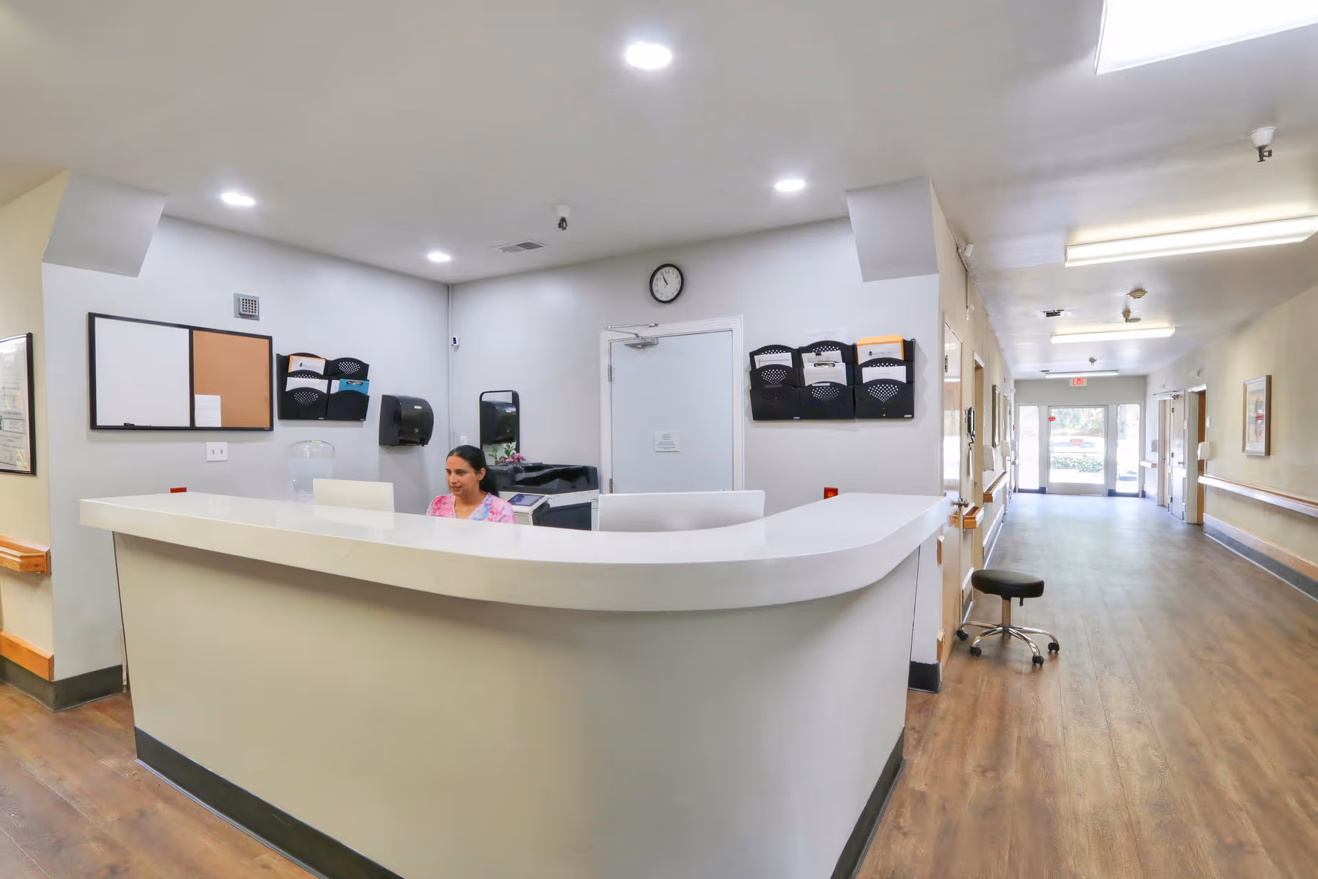 Reception desk in a senior healthcare facility lobby with a staff member seated behind it and a long hallway leading to an exit.