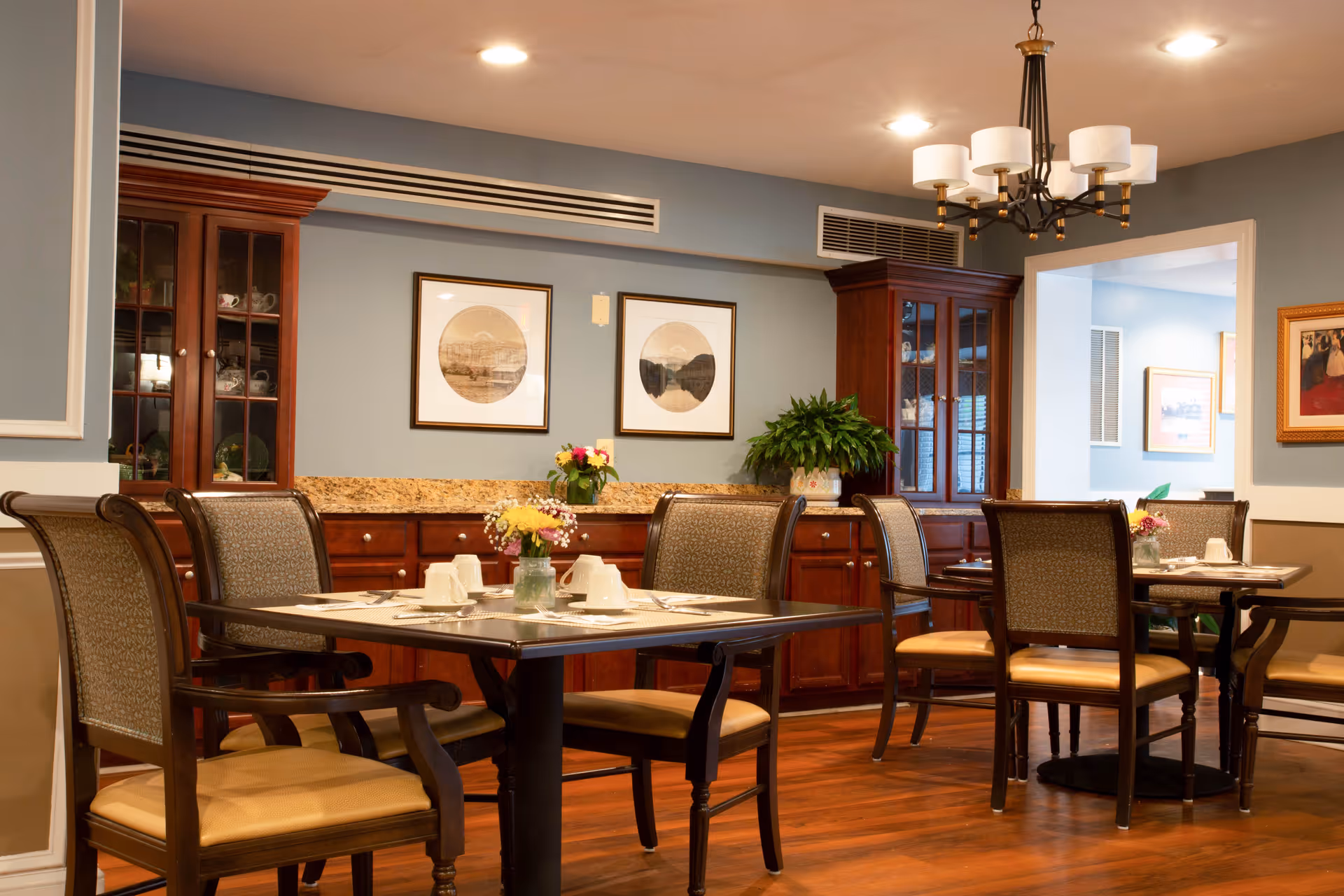 A cozy dining room with wooden floors, several tables set with placemats, cups, and small flower arrangements. The room features wooden chairs with cushioned seats, a wooden cabinet with glass doors displaying dishes, framed artwork on the walls, and a chandelier hanging from the ceiling.