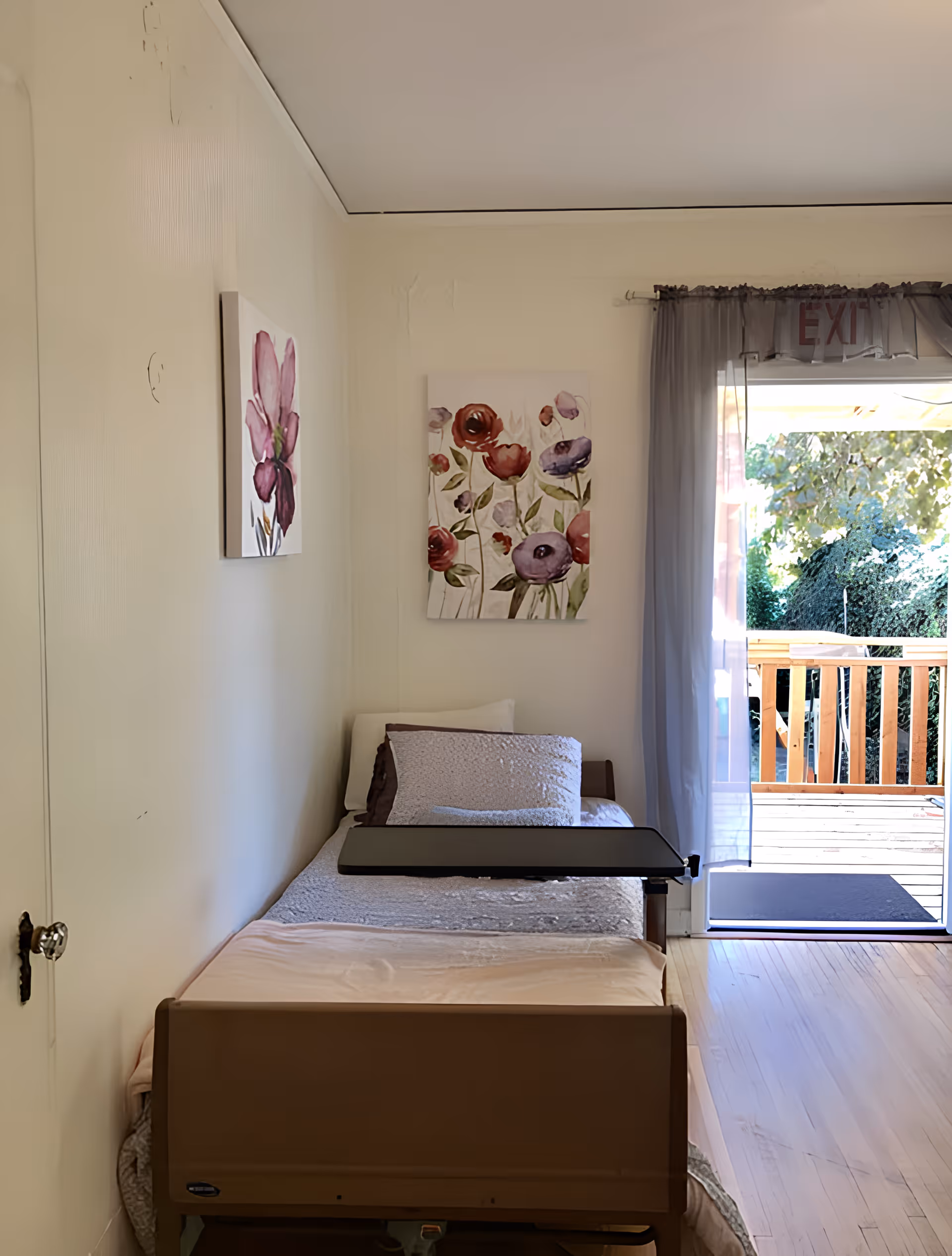 A small bedroom with a single hospital-style bed positioned against a cream-colored wall. The bed has a light pink blanket, a gray textured pillow, and a white pillow. Above the bed are two floral paintings with pink and red flowers. To the right, there is a sliding glass door with sheer gray curtains, leading to an outdoor wooden deck with greenery visible outside. An exit sign is partially visible above the door.