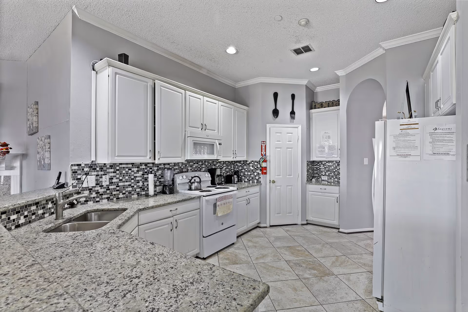 A modern kitchen with white cabinets, granite countertops, and a tiled floor. The kitchen features a white stove with a kettle, a blender, a coffee maker, and a double sink. The backsplash is made of small square tiles in shades of gray and black. There is a white refrigerator with papers attached to it, a pantry door, and decorative oversized fork and spoon wall hangings. The ceiling has recessed lighting.