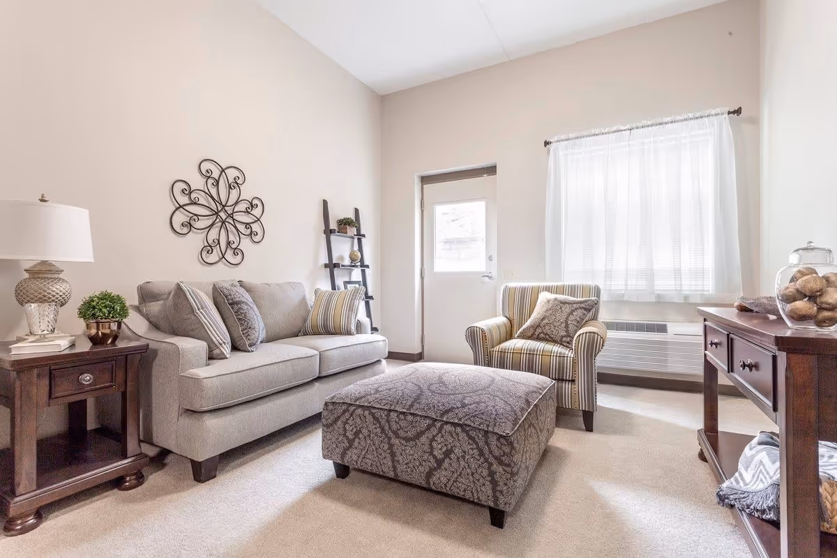 A cozy living room with a light gray sofa adorned with multiple cushions, a patterned ottoman in front, a striped armchair with a decorative pillow, a wooden side table with a lamp and a small plant, a wooden console table with decorative items, and a window with sheer white curtains letting in natural light.