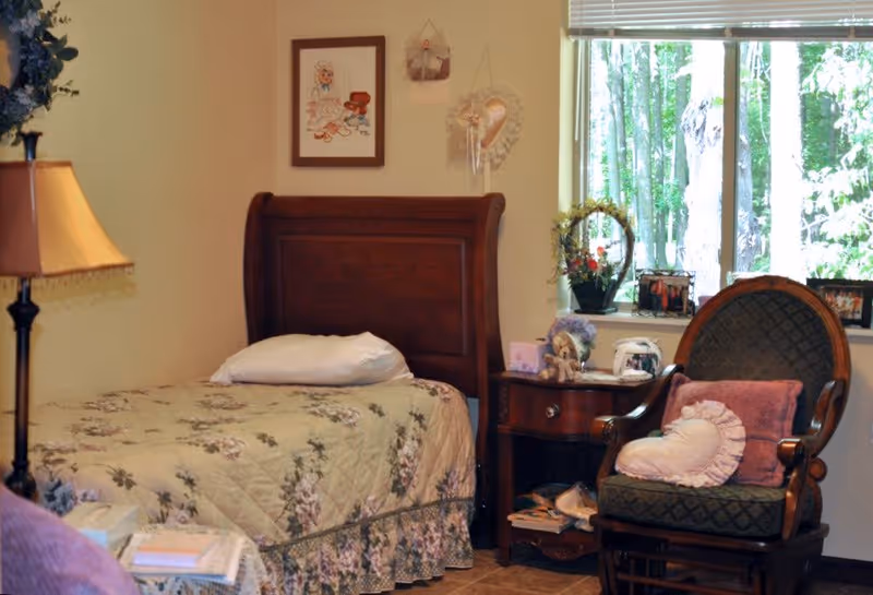 A cozy bedroom with a single wooden bed covered in a floral quilt and a white pillow. Next to the bed is a wooden nightstand with various decorative items and a small lamp. A cushioned armchair with a heart-shaped pillow is positioned near a window showing green trees outside. The walls have framed artwork and hanging decorations.