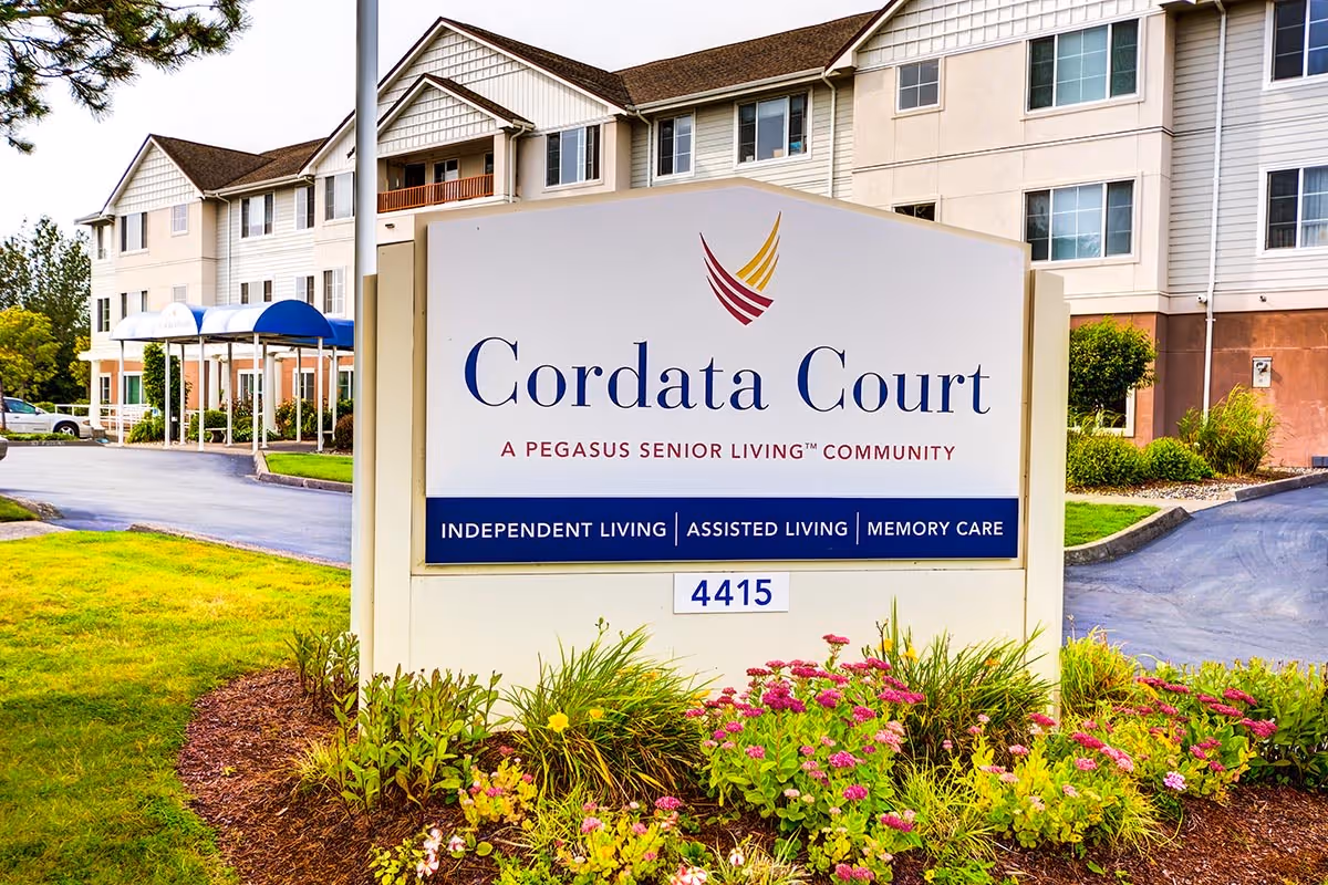 Outdoor view of the entrance sign for Cordata Court, a Pegasus Senior Living community offering independent living, assisted living, and memory care. The sign is surrounded by flowers and greenery, with a multi-story building and a driveway in the background.