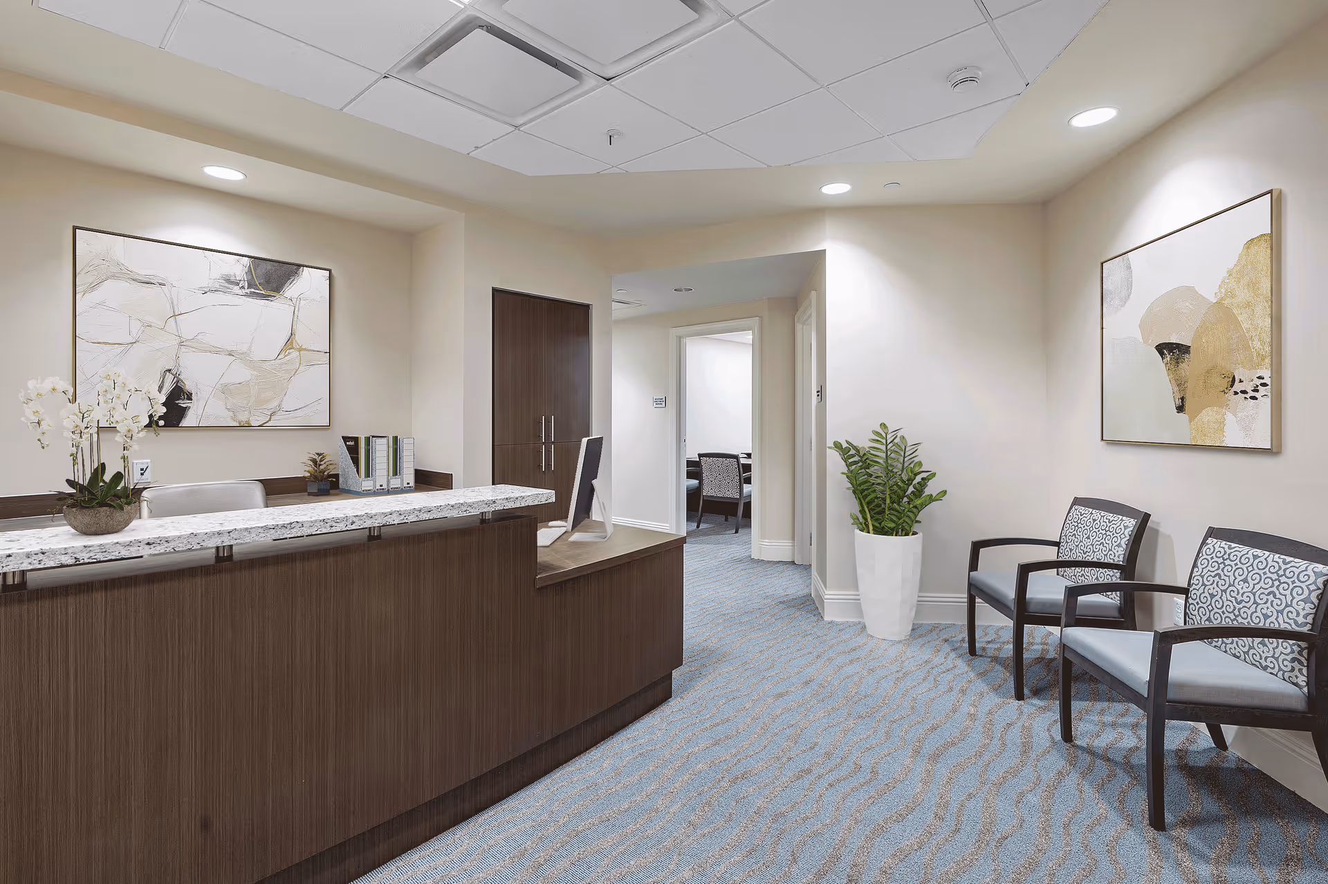 Reception area with a wooden front desk topped with a white and gray marble surface, a computer monitor, and a potted orchid plant. Two patterned chairs are placed against the wall on the right, next to a large potted green plant. The walls are decorated with abstract artwork, and the floor is covered with a blue patterned carpet. There is a hallway leading to other rooms in the background.