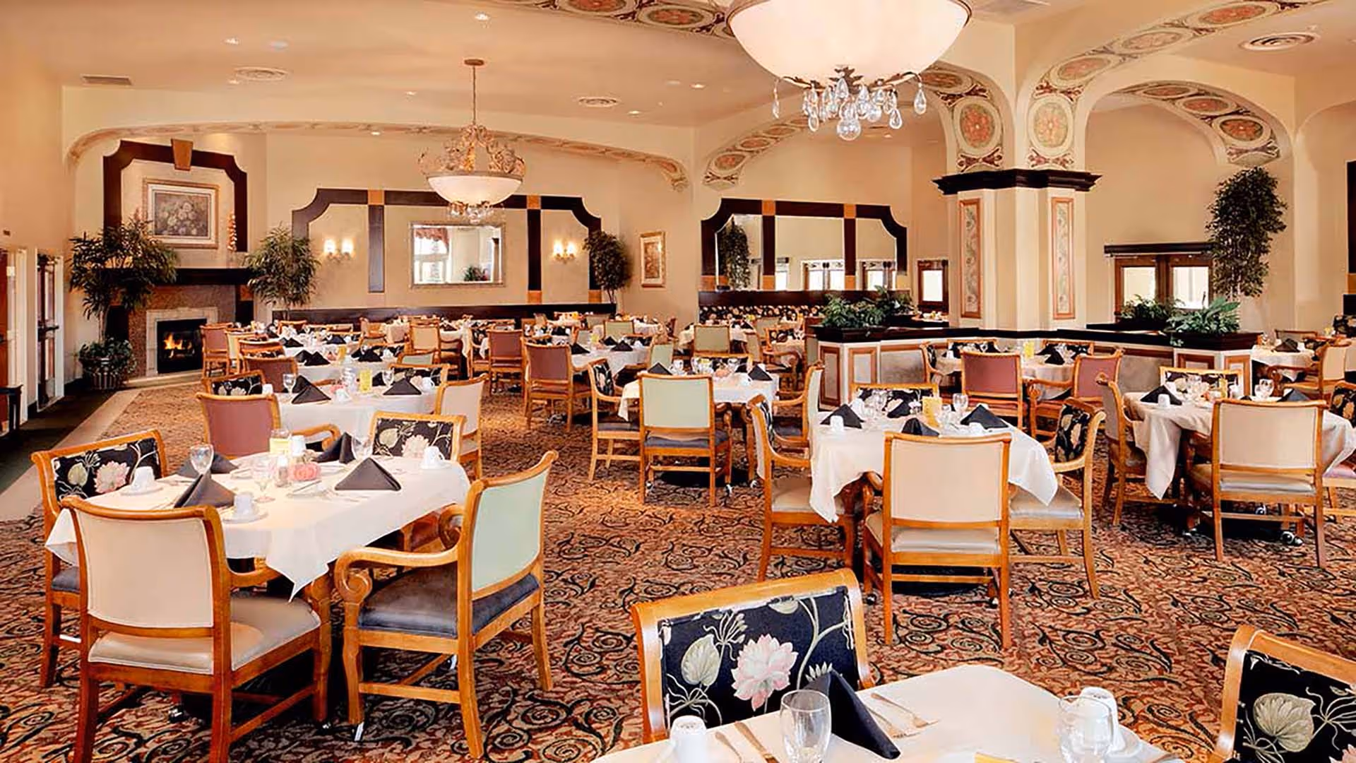 A spacious dining room with multiple tables covered in white tablecloths, each set with black napkins, glassware, and utensils. The room features patterned carpet, decorative ceiling arches, chandeliers, large mirrors on the walls, and potted plants. There is a fireplace in the back corner.
