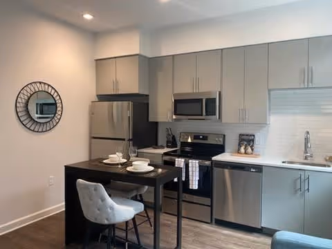 Modern kitchenette with stainless steel appliances, gray cabinets, a small dark dining island set for two and a round wall mirror.