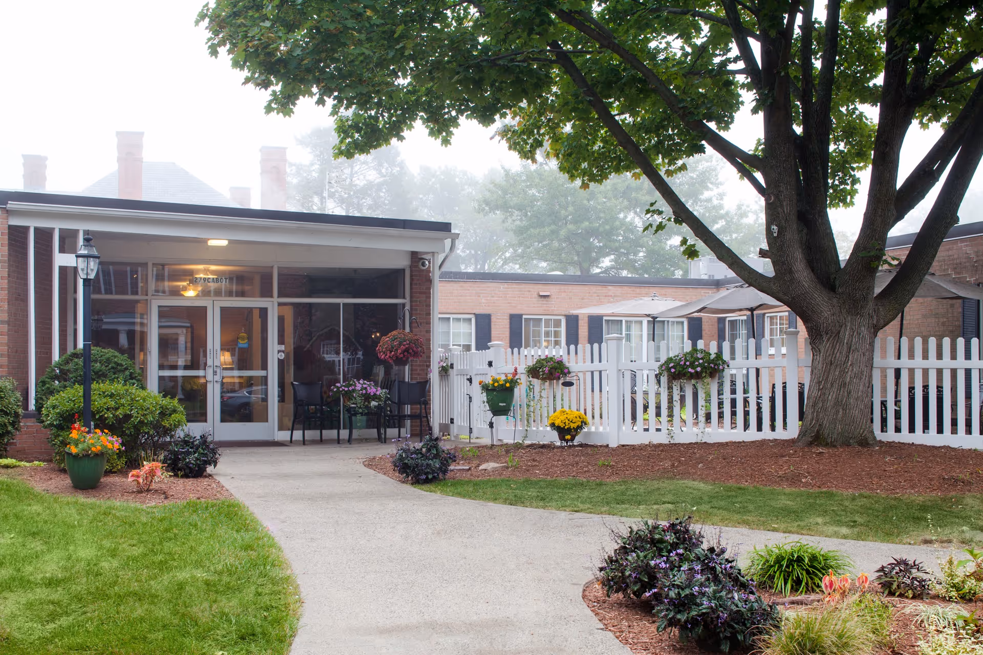 Entrance to Renaissance Manor on Cabot showing a brick building with glass doors, a white picket fence, outdoor seating with chairs and tables, and landscaped garden beds with flowers and shrubs under a large tree.