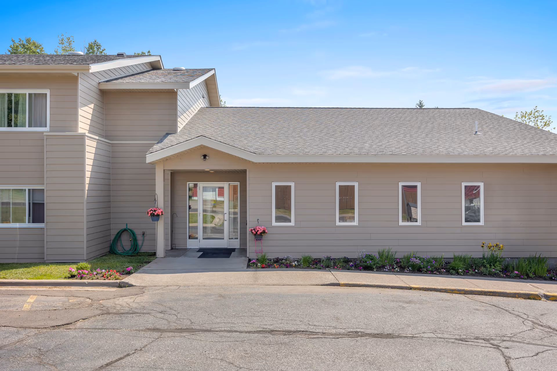 Exterior front entrance of a beige senior living building with a covered doorway, double glass doors, windows, and flowerbeds.