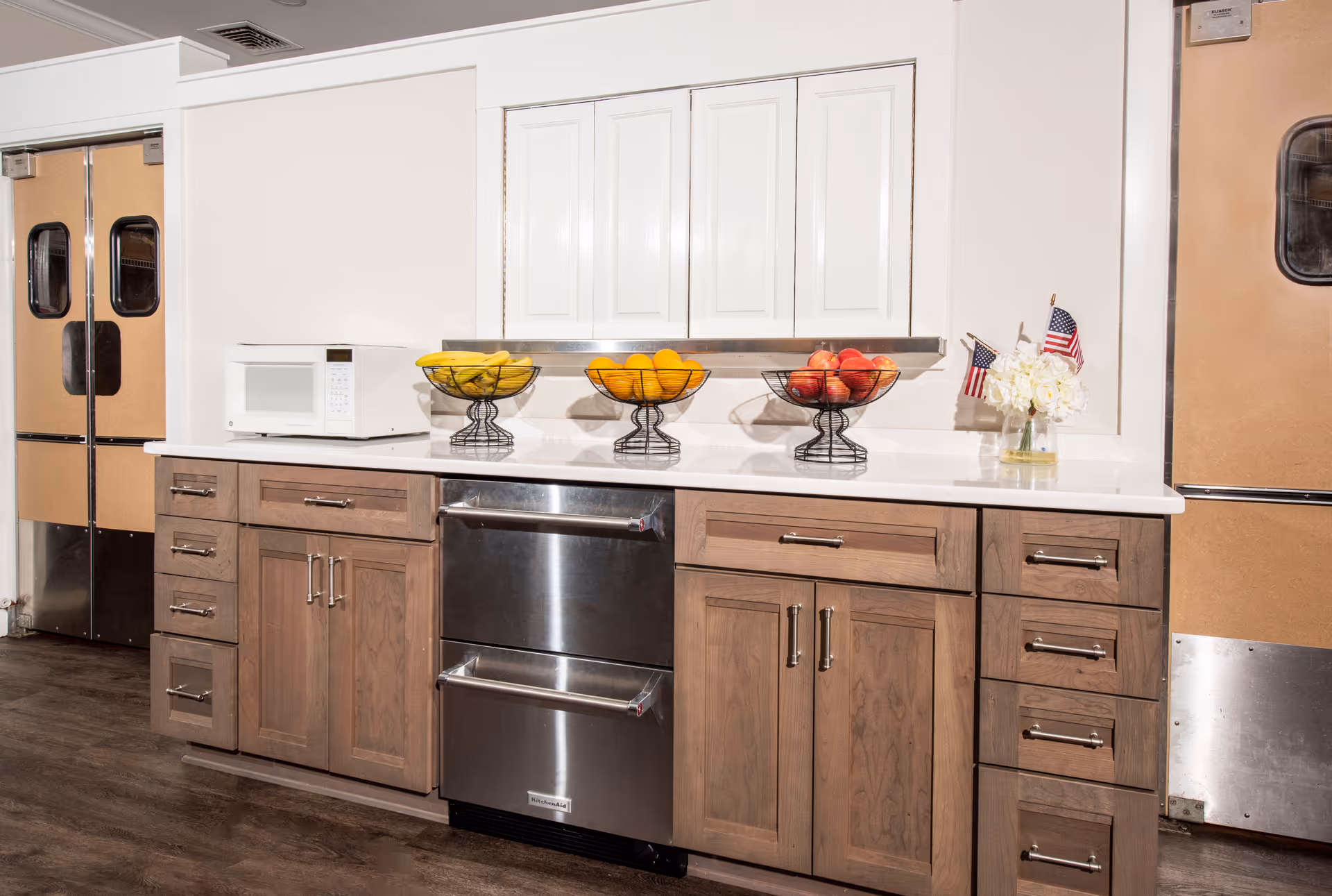 A kitchen area with wooden lower cabinets and white upper cabinets. On the countertop, there is a white microwave, three wire fruit baskets containing bananas, oranges, and apples, and a small vase with white flowers and two small American flags. The floor is dark wood, and there are two metal double doors on either side of the kitchen area.