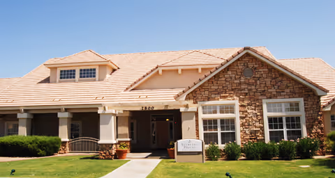 Exterior view of a single-story senior living facility building with a beige tiled roof, stone and stucco walls, large windows, and a well-maintained lawn with shrubs. A sidewalk leads to the entrance under a covered porch.