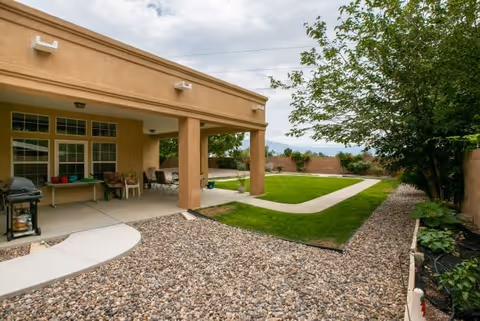 Outdoor patio area of a senior living facility with a covered seating area featuring chairs and a table, a barbecue grill, a pathway bordered by gravel and green grass, and trees along a brick wall under a cloudy sky.