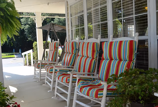A row of white wooden rocking chairs with colorful striped cushions on a covered porch. The porch has large windows with white blinds and some green plants nearby. In the background, there is a swing and greenery.