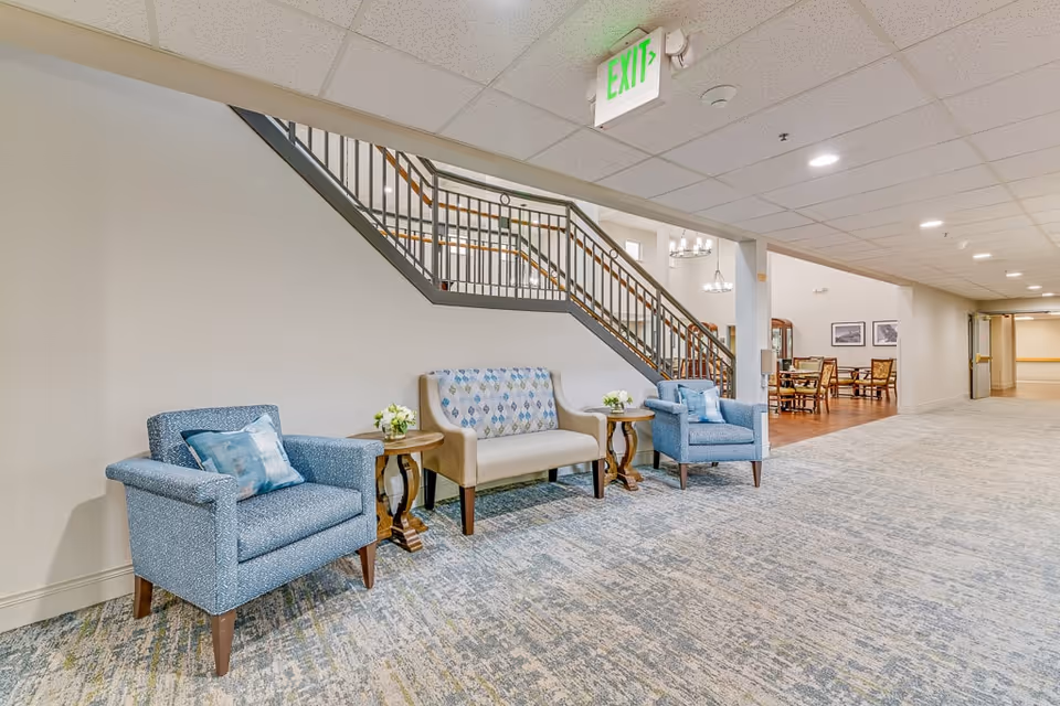 A seating area in a senior living facility hallway featuring two blue armchairs and a beige loveseat with patterned upholstery, separated by two small wooden side tables with flower arrangements. Behind the seating area is a staircase with a metal railing. The hallway extends to a dining area with tables and chairs visible in the background. The ceiling has recessed lighting and an illuminated exit sign.