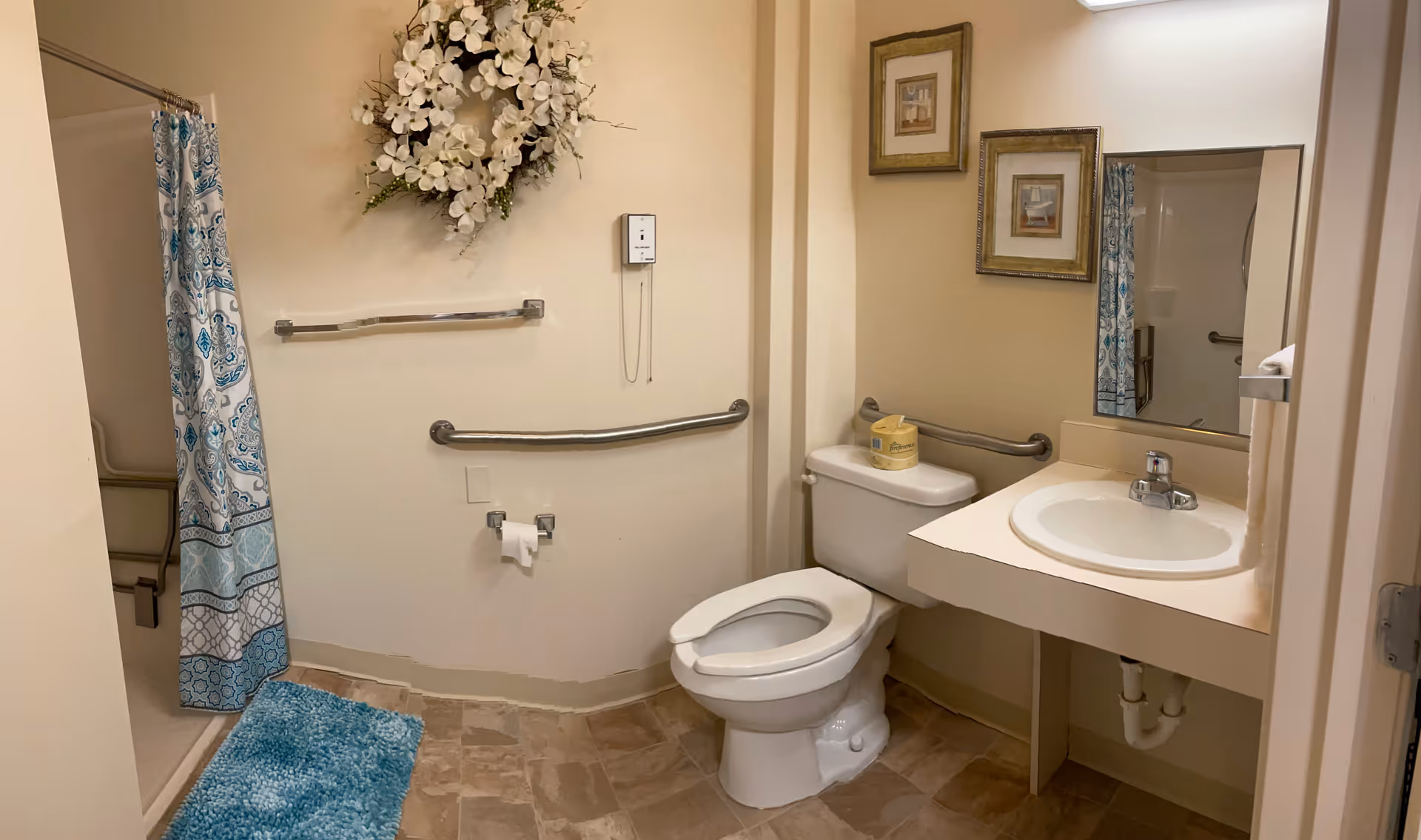 A clean and accessible bathroom with a toilet, sink with a mirror above it, and a shower with a blue and white patterned curtain. There are safety grab bars near the toilet and in the shower area. A blue bath mat is placed on the floor, and a decorative white flower arrangement hangs on the wall above the towel rack. Two framed pictures are hung on the wall next to the mirror.