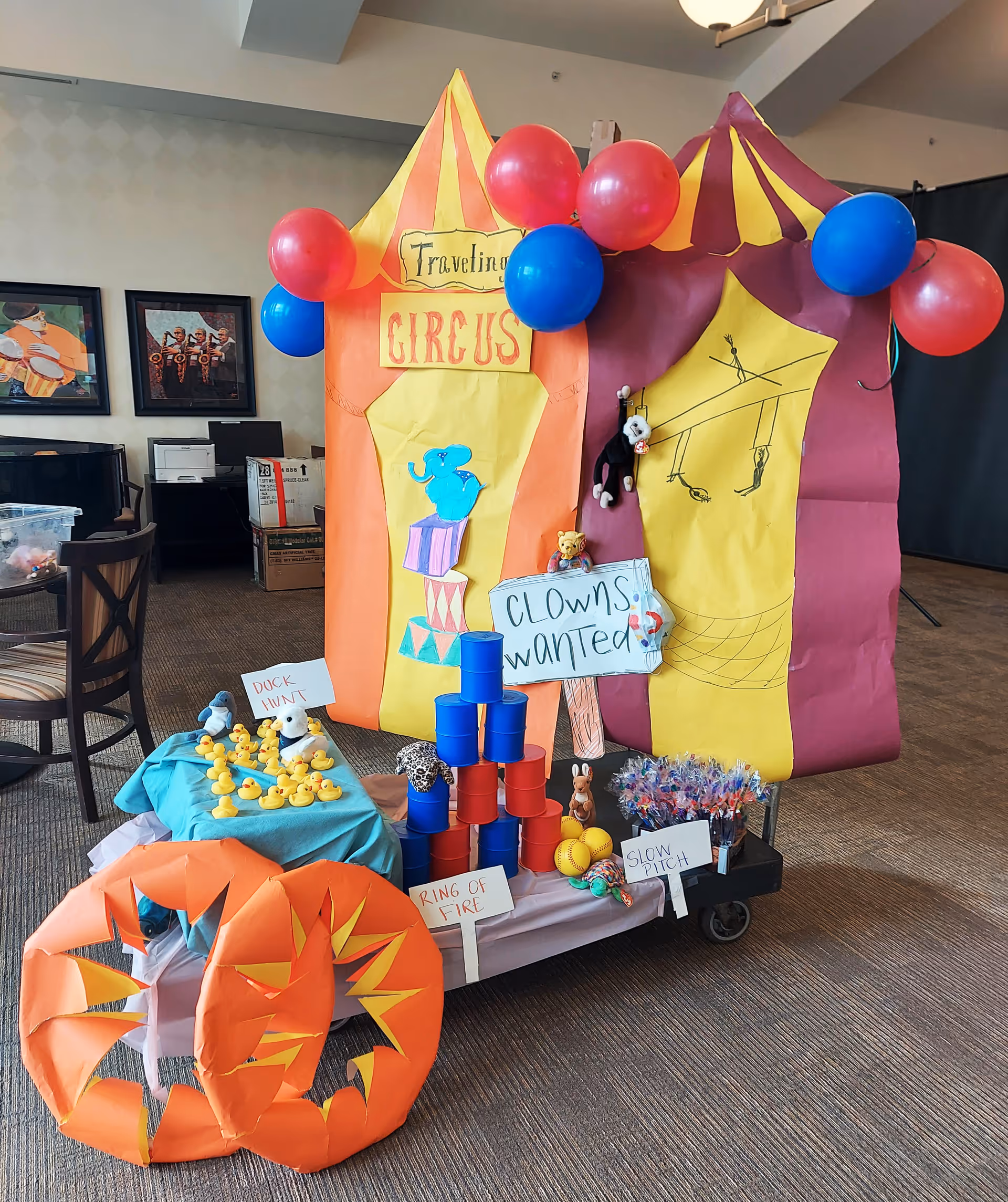 A colorful indoor circus-themed display with a large paper circus tent decorated with red and blue balloons. The tent has signs reading 'Traveling Circus' and 'Clowns Wanted.' In front of the tent are various carnival game props including a 'Duck Hunt' with rubber ducks, a 'Ring of Fire' with stacked red and blue cans, and a 'Slow Pitch' game with softballs and lollipops. The setting appears to be a common area with chairs, framed pictures, and boxes in the background.