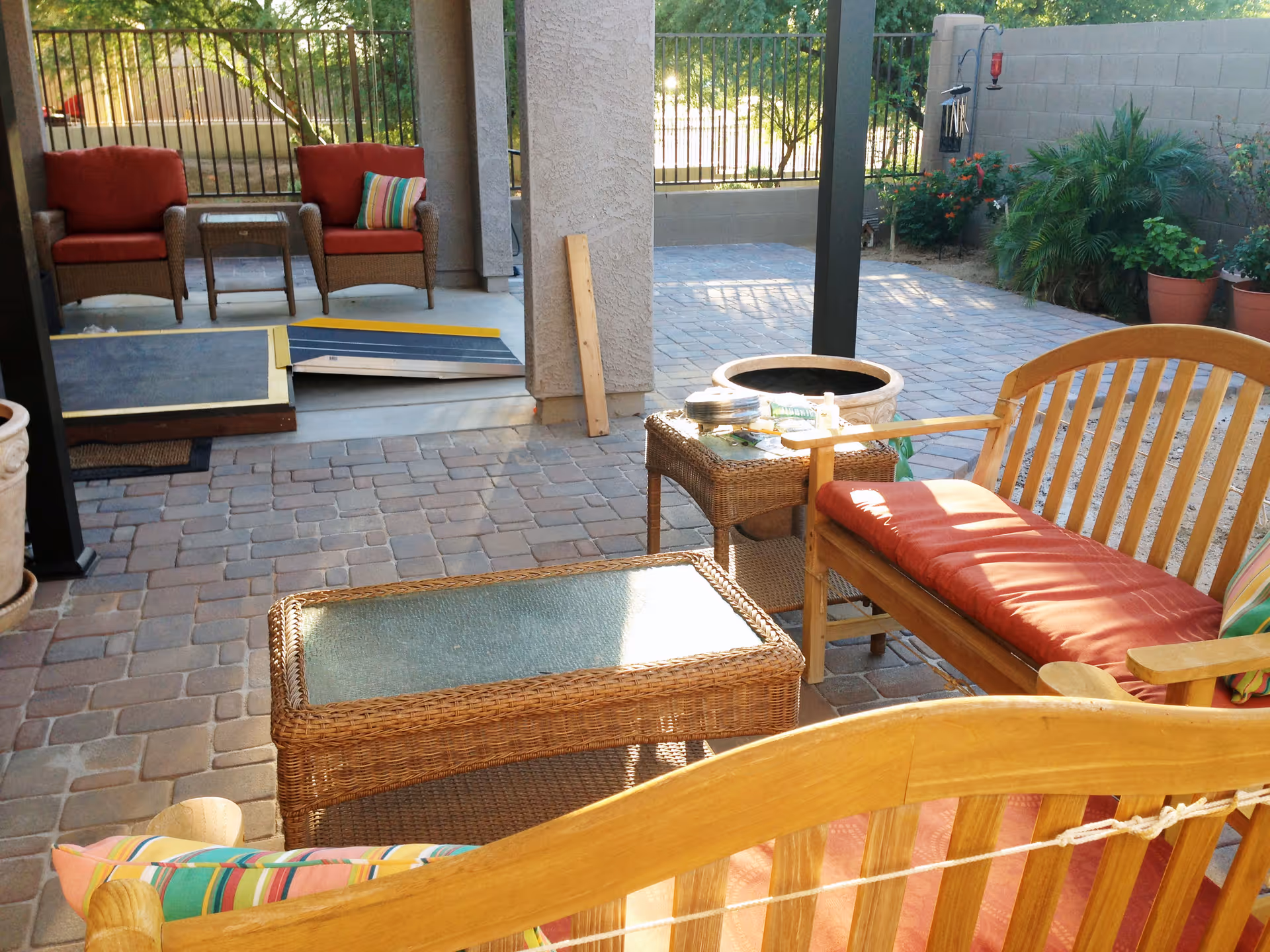 Outdoor patio area with wicker and wooden furniture including chairs and a bench with red cushions and colorful striped pillows. There is a glass-top wicker coffee table and a small wicker side table with items on it. The patio has stone pavers and is partially shaded by a covered structure. In the background, there is a ramp leading to a door, a fence, and some plants in pots and garden beds.