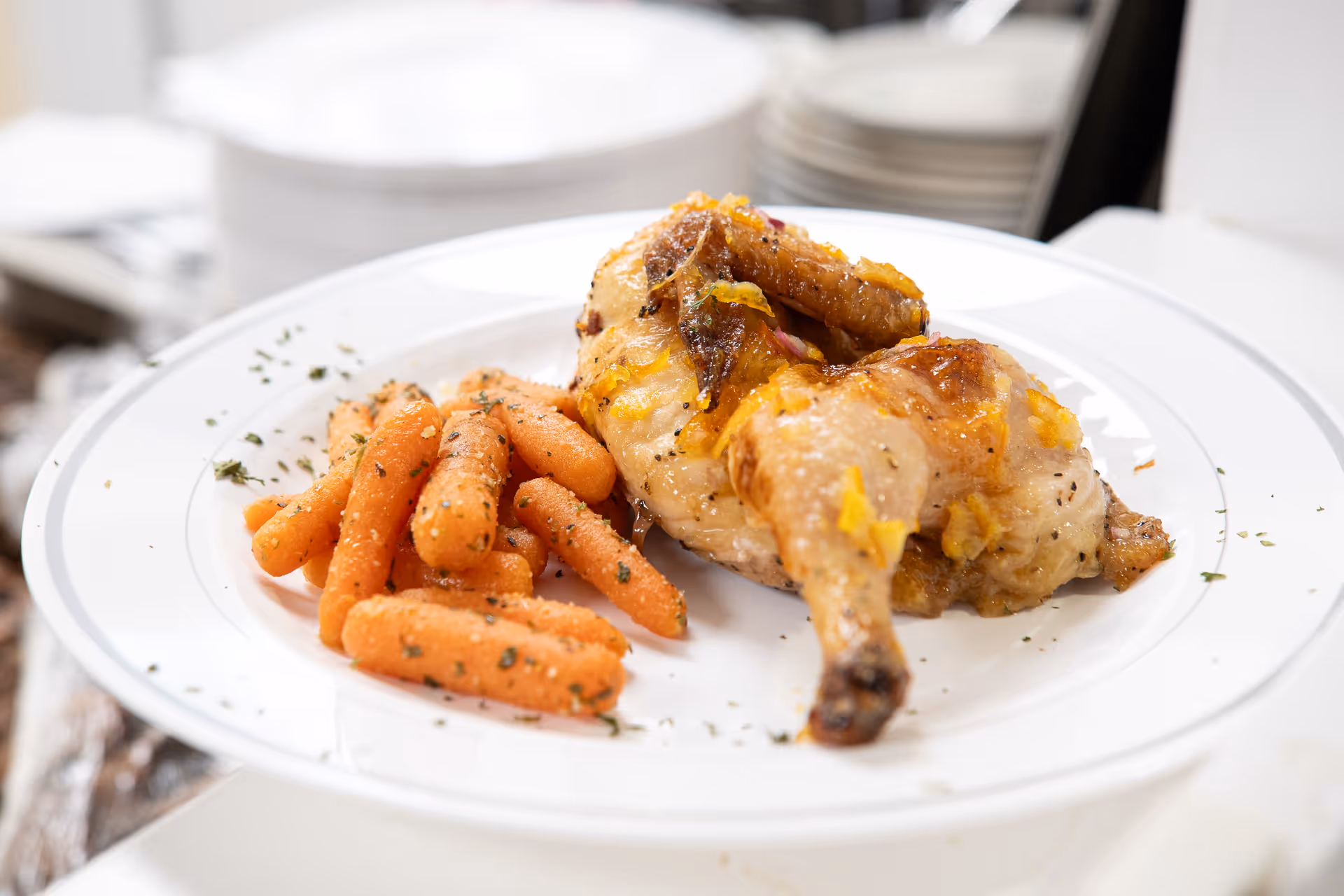 A white plate with a serving of roasted chicken and cooked baby carrots garnished with herbs, with stacked white plates blurred in the background.