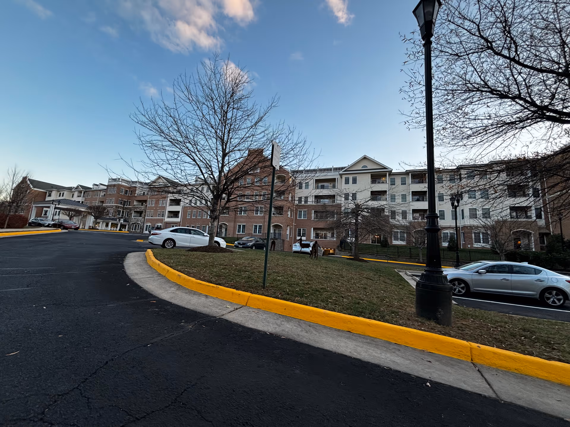 Exterior view of a multi-story senior living facility building with a parking lot in front. The building has a combination of brick and light-colored siding with multiple balconies. Leafless trees and a streetlamp are visible in the foreground under a partly cloudy sky.