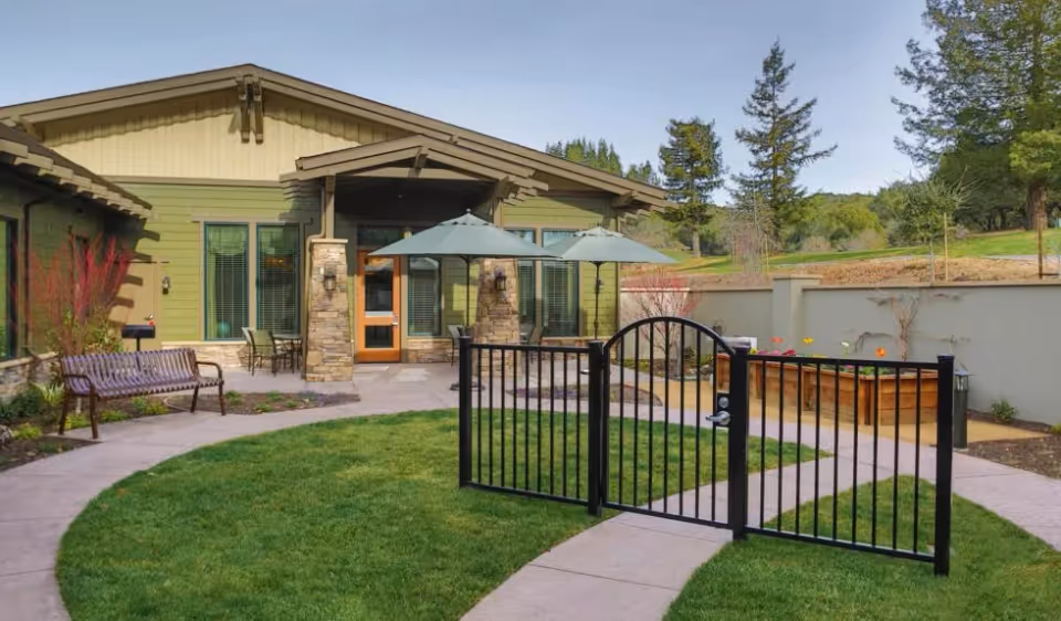 Outdoor courtyard area of a senior living facility with a green lawn, a black metal gate, a paved walkway, a wooden bench, two patio tables with umbrellas, and a green building with stone pillars in the background surrounded by trees and a low wall.