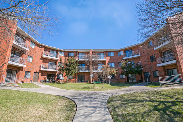Three-story red brick apartment building forming a U around a grassy courtyard with walkways and balconies under a clear blue sky.