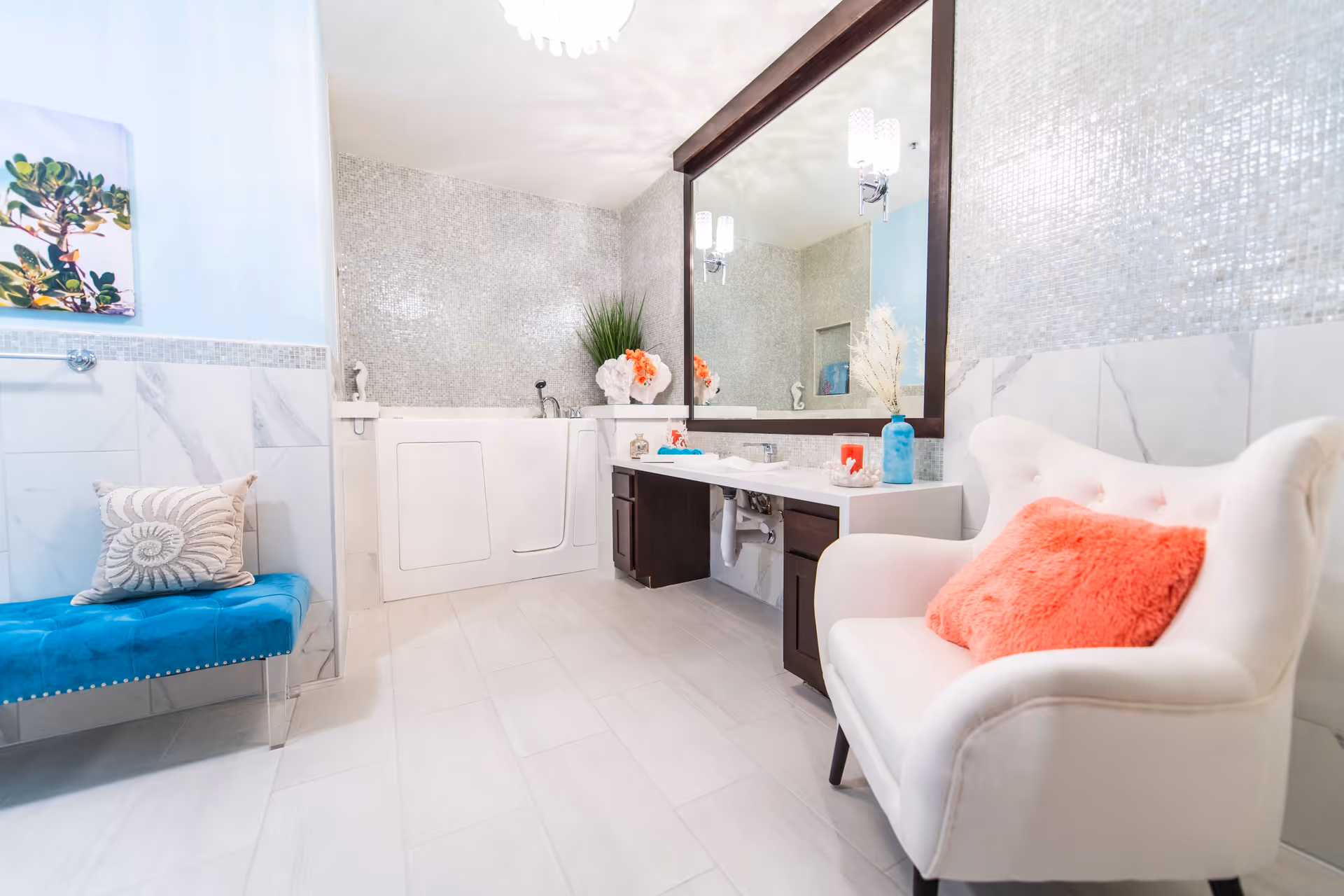 Bright and spacious bathroom with a walk-in bathtub, large mirror above a double sink vanity with dark wood cabinets, a white armchair with a coral pillow, a blue bench with a decorative pillow, and a wall painting. The walls have a mix of light blue paint, marble tiles, and small mosaic tiles.