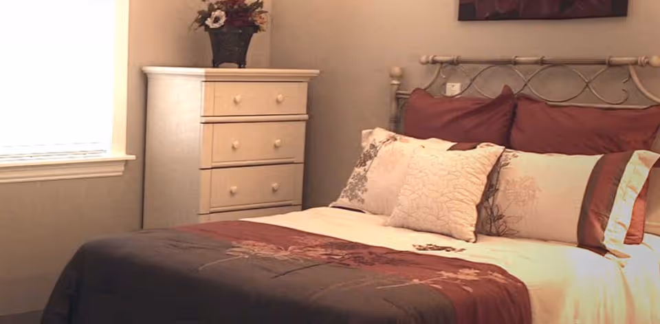A neatly made bed with multiple pillows and a decorative bedspread in a softly lit bedroom. Next to the bed is a white chest of drawers with a flower arrangement on top, and a window with blinds partially open letting in natural light.