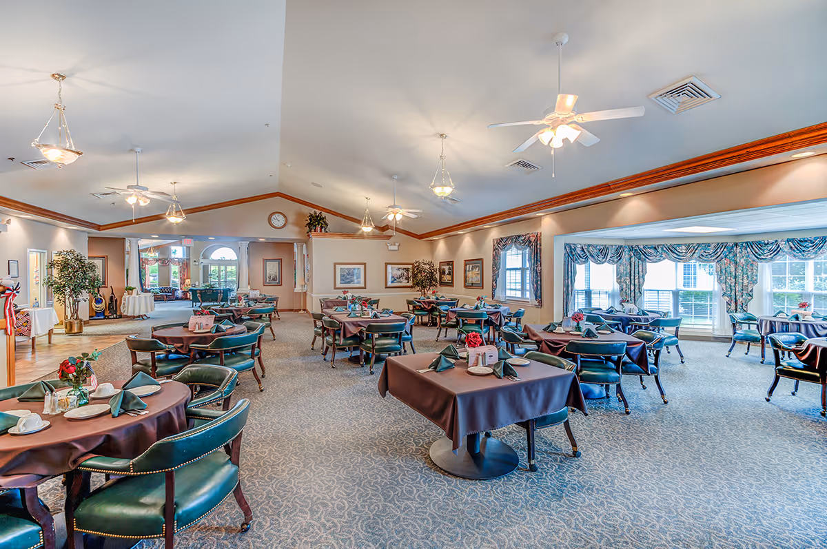 Spacious dining room with multiple tables covered in dark tablecloths, each set with green folded napkins, plates, cups, and small flower arrangements. The room features carpeted floors, ceiling fans, hanging light fixtures, large windows with floral curtains, and framed artwork on the walls.